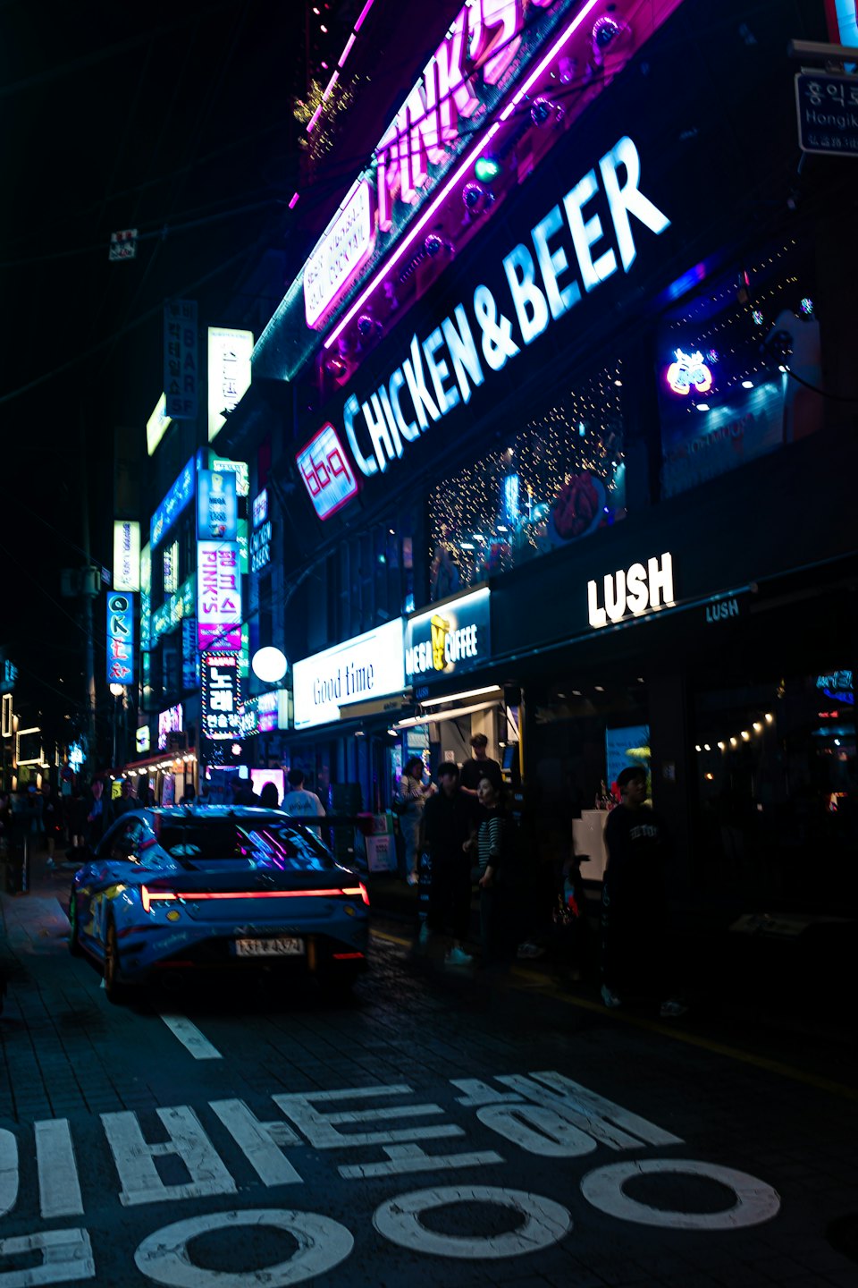 Un coche deportivo azul circula por una calle de ciudad iluminada con luces de neón por la noche.