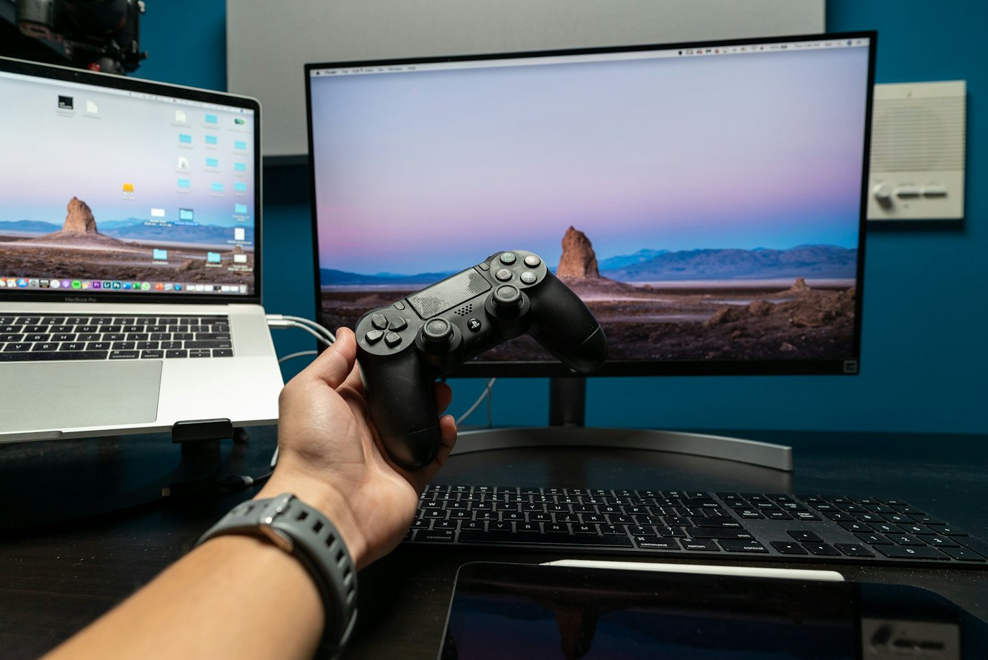 Mano sujetando un mando de videojuegos cerca de un portátil y un monitor.